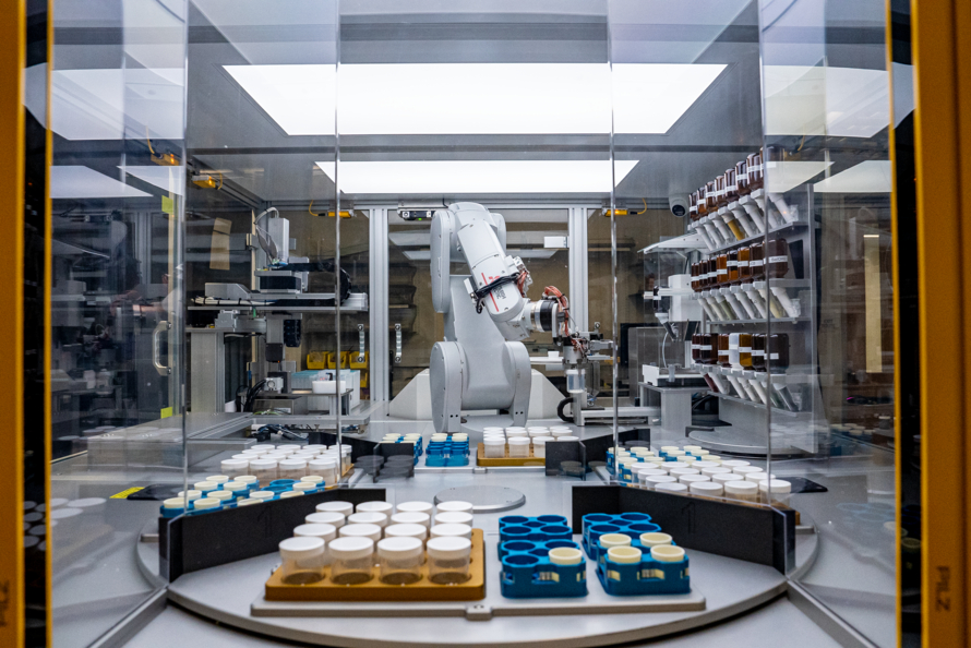 A robot inside a glass enclosure with bottles of chemicals and sample jars